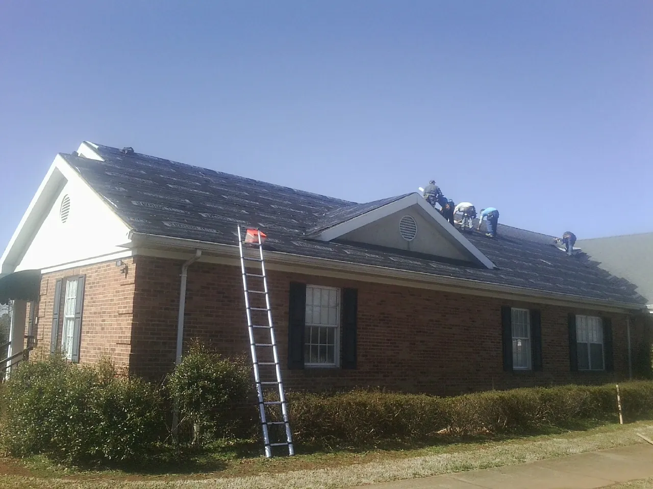 Crew tearing shingles off a brick ranch roof with a ladder set — Emergency Roof Repair work by our crew in Mc Kenney