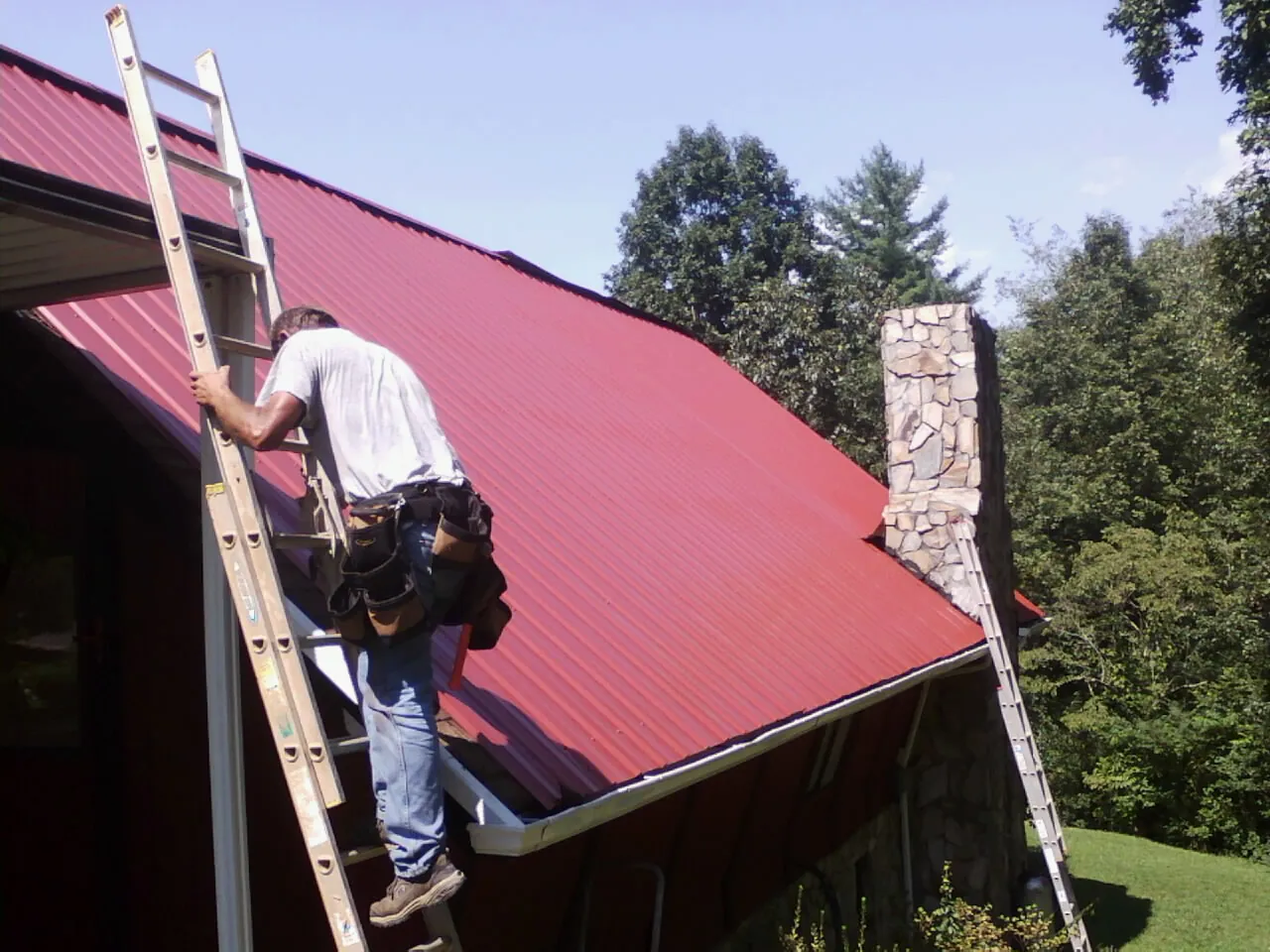 Roofer on a ladder beside a red metal roof and stone chimney — Soffit & Fascia Repair by Roofing Brothers in Mc Kenney
